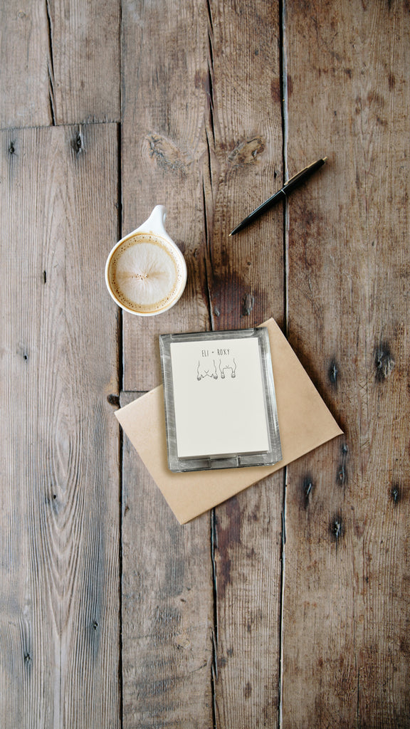 Cup of coffee, notepad with acrylic holder and sploot designs, and pen on a wooden surface
