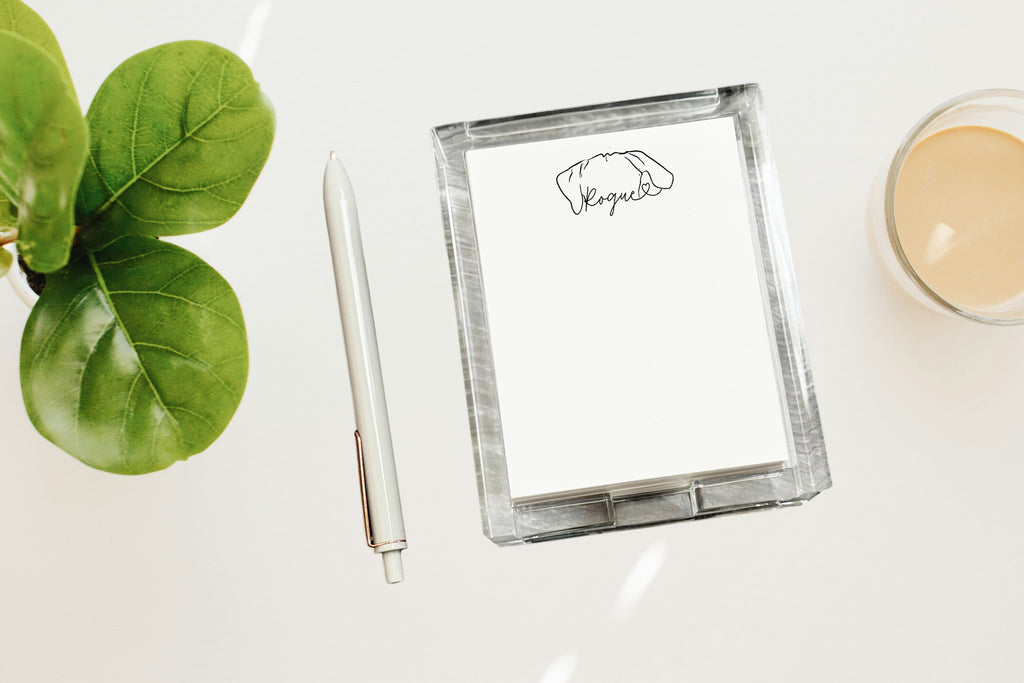 Clear acrylic notepad holder with custom pet ears with a logo, pen, and plant on a white surface
