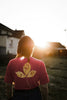 Person wearing a red shirt with a leaf design with paw prints, standing in a field with the sun setting behind them.