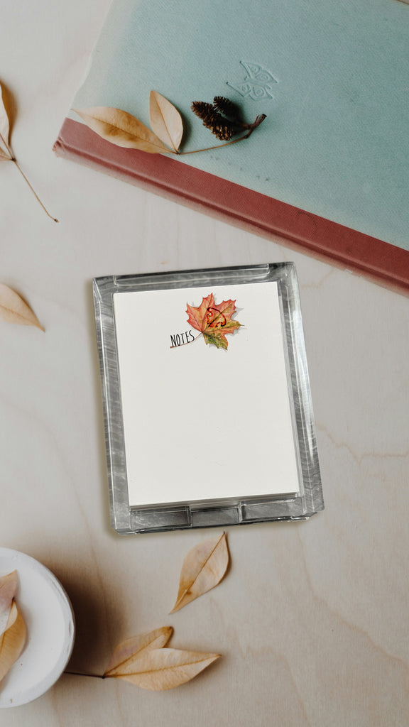 Small notepad with nose print on leaf design imprinted with "notes" wording on a reflective surface, with a book and dried leaves in the background.
