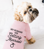 A small fluffy dog sitting on a white bed wearing a light pink long-sleeve shirt with the black printed text 'MY HUMANS ARE Getting Married' and a ring graphic.