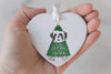 Heart-shaped ornament with a dog in a Christmas tree costume and Tis the Season to Sparkle text, held in a hand against a light background.