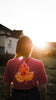 Person wearing a crimson Comfort Colors shirt with a leaf design printed with a paw, standing in a field with a sunset in the background.