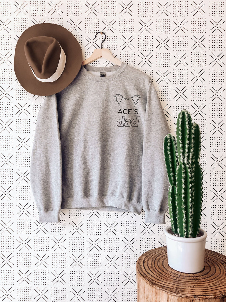Personalized Pocket Dog Dad, Cat Dad, or Other Pet's Ears Outline Tattoo Inspired Crew Neck Unisex Sweatshirt in Heather Grey - Personalized with Ace's Dad - Sweatshirt hanging up next to a hat and a cactus